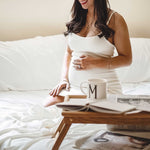 pregnant woman in bed on ivory bamboo bed sheets with cup of team and smiling