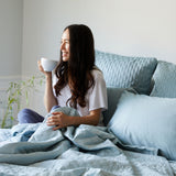 dark haired lady drinking coffee in bed in sky blue coverlet and pillows lounging in bed