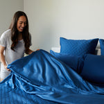 smiling lady holding onto a flat feet of dark indigo blue bamboo bed sheets making the bed