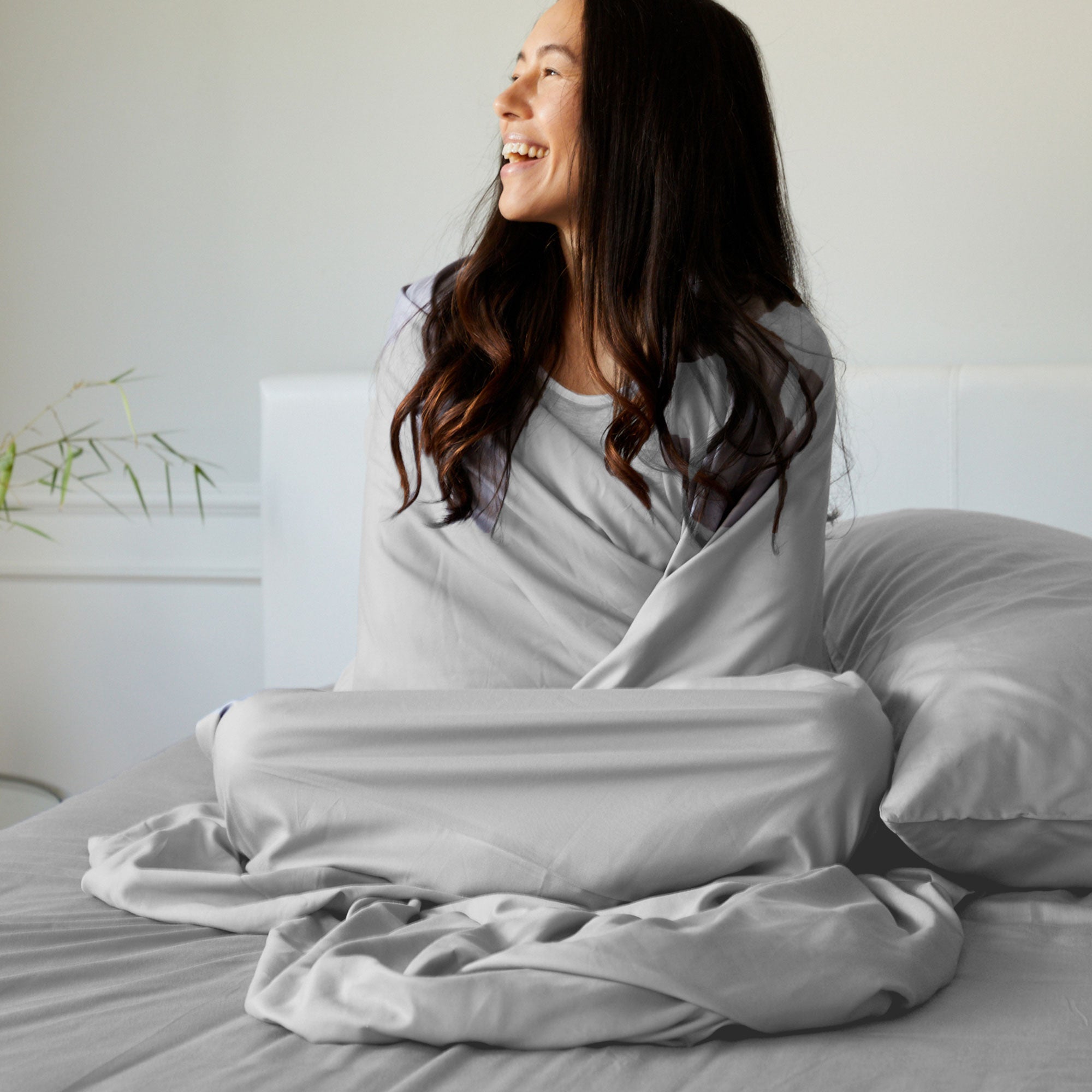 dark haired woman laughing while wrapped in light gray bamboo bed sheets on bed