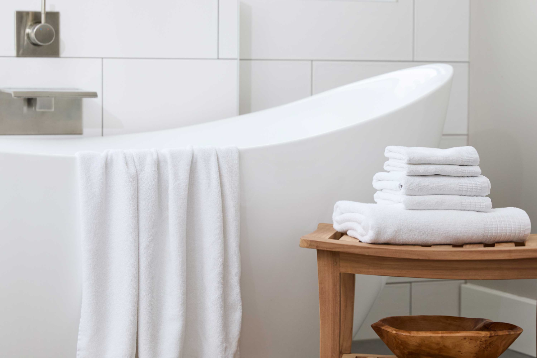 cloud white bamboo towels draped over a white bathtub and 5 more white bamboo towels stacked on a bamboo stool
