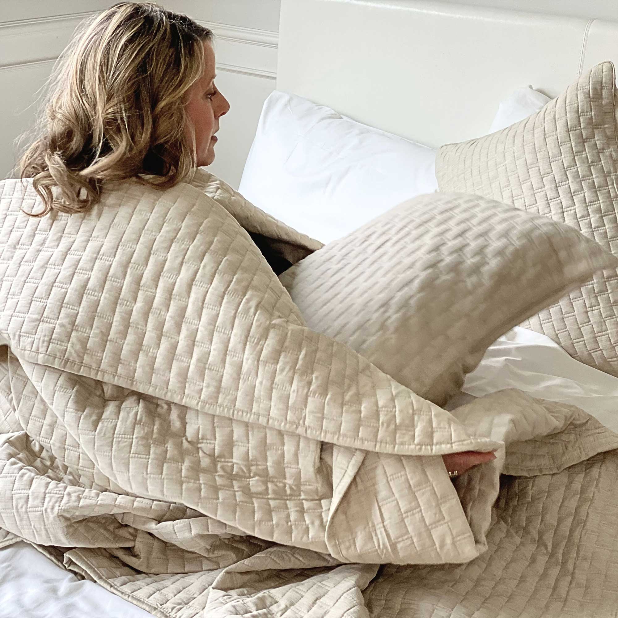 woman wrapped in a sand beige melange bamboo quilted coverlet on a bed looking cozy