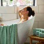 pretty woman with dark hair lounging in a modern white bathtub  with seaglass green bamboo towels draped over tub