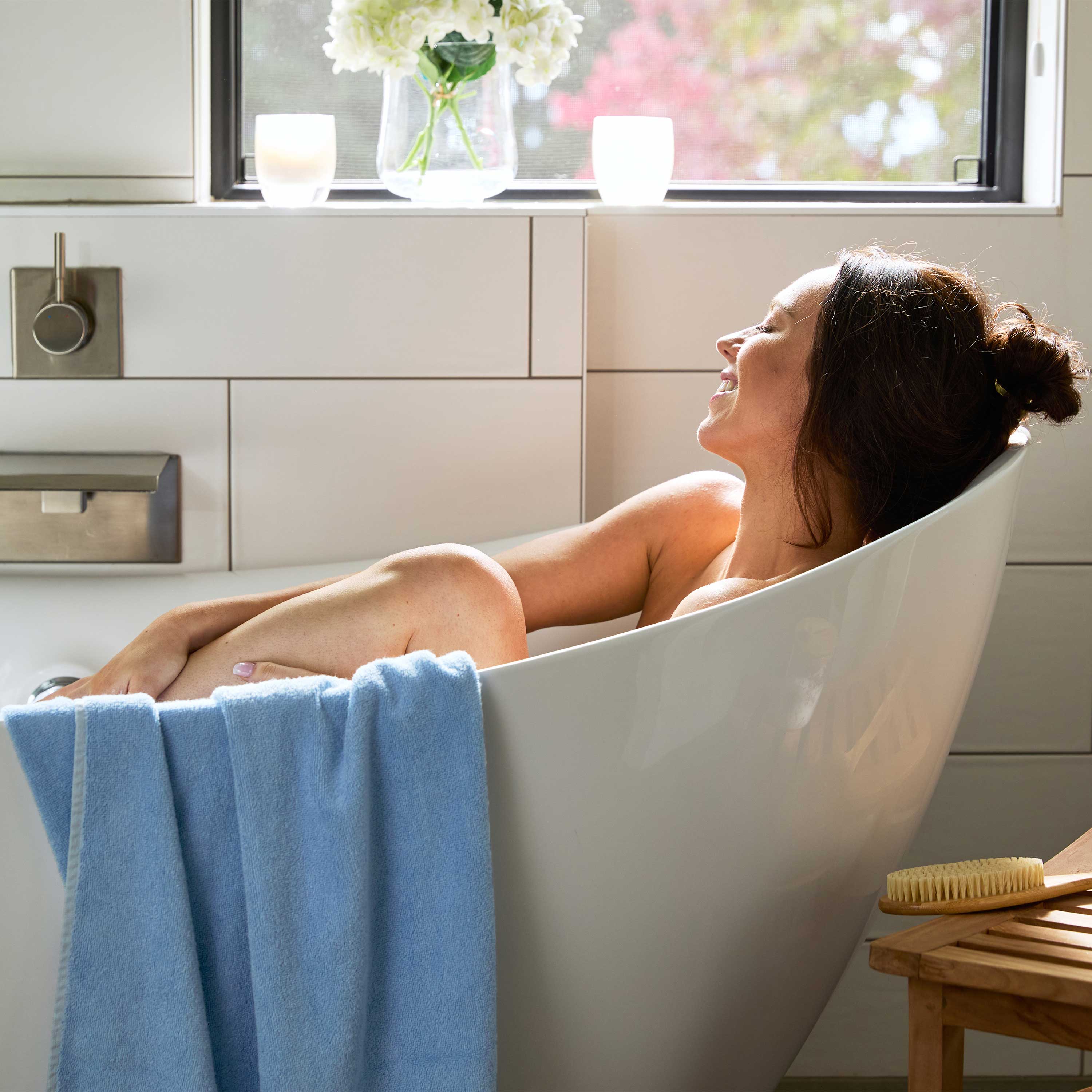 woman soaking in bathtub with ocean blue bamboo towel draped over the tub in modern bathroom and window