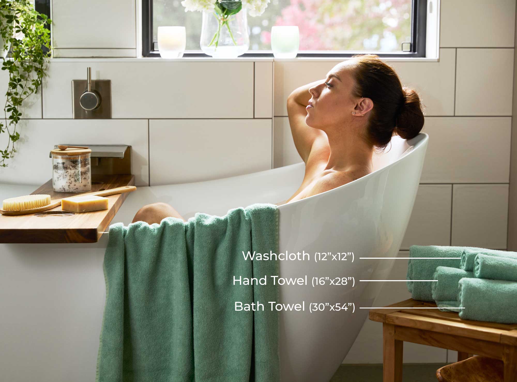 woman soaking in white bathtub with seaglass green towels draped over tub and rolled on wood stool