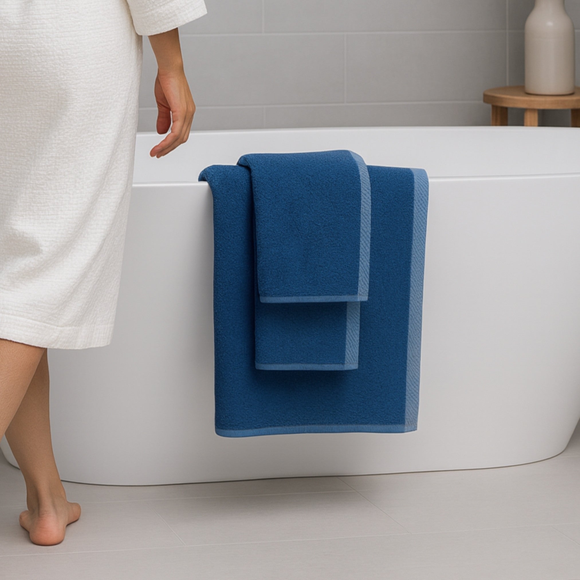 Person in a white robe standing next to a bathtub with blue towels draped over it, in a bathroom setting.