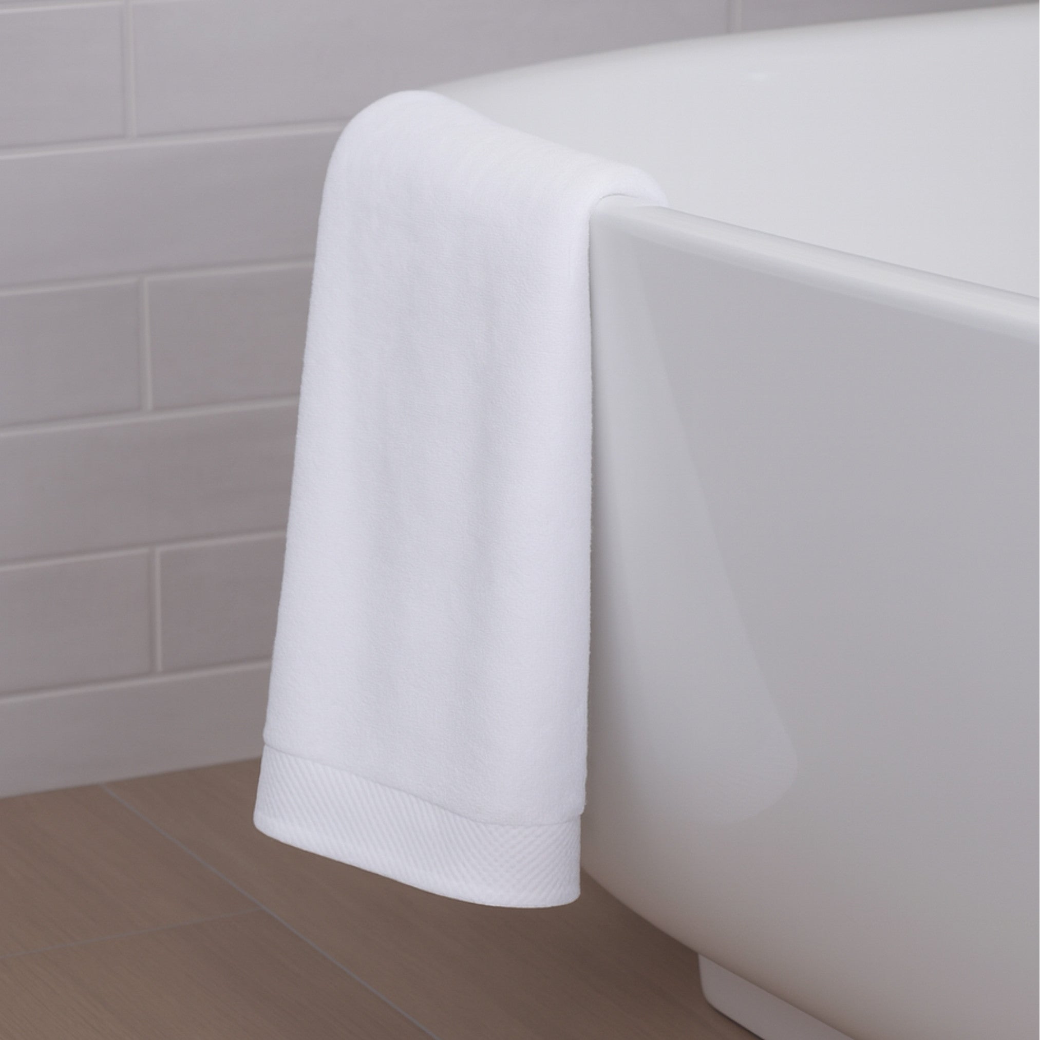 White bath towel draped over a white bathtub against a tiled bathroom wall.