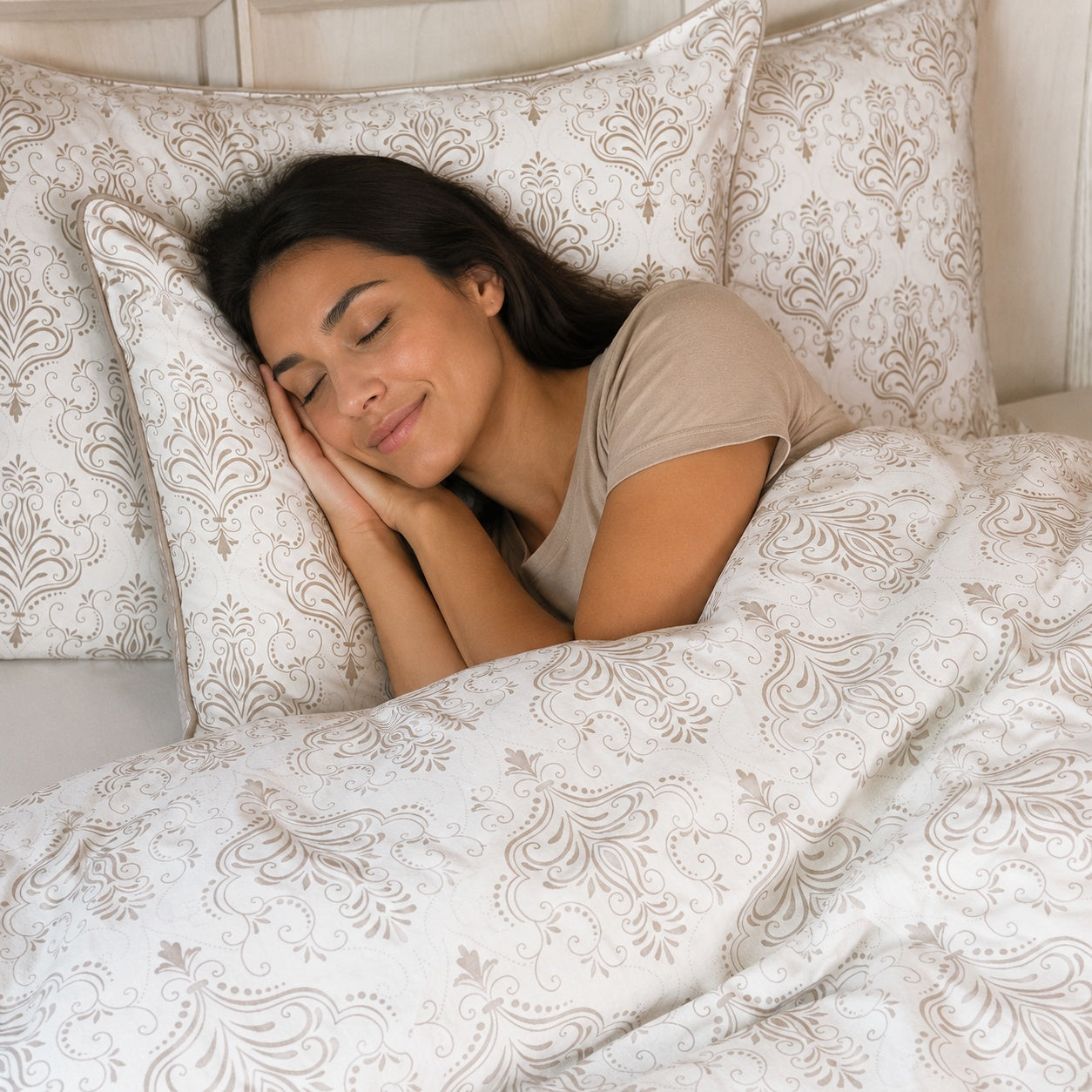 A woman sleeping peacefully in bed, covered with a white duvet featuring a beige damask pattern, with matching pillows behind her.