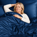 A woman with blonde hair lies in bed under a shiny navy blue duvet, with matching navy pillows behind her. She is wearing a white long-sleeve top.