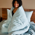 woman wrapped up in sky blue duvet cover on a wood bed and tan walls