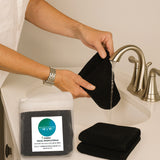 Person holding a black washcloth over a sink with a package of washcloths labeled 'Bed Voyage' in the foreground.