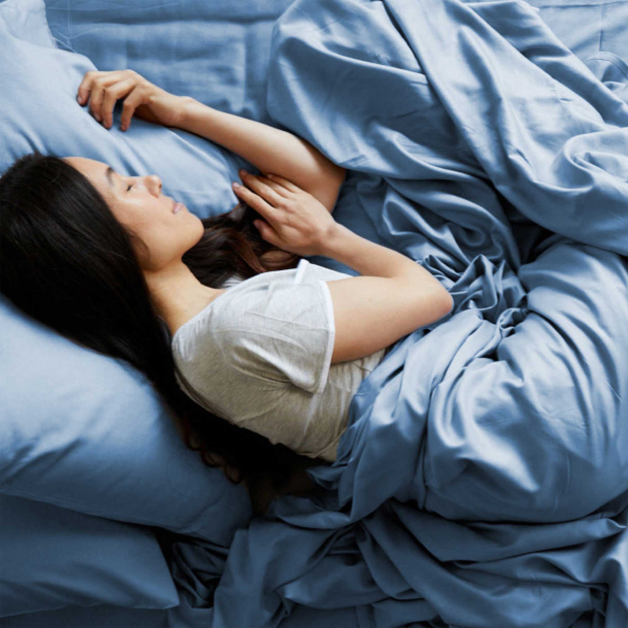 Woman lying in bed under blue sheets and pillows, wearing a white shirt.