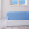 A blue fitted sheet on a mattress with a white base, placed in a room with a light wall and a window with a view of the ocean.