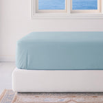 Light blue fitted sheet on a white bed in a room with a window.
