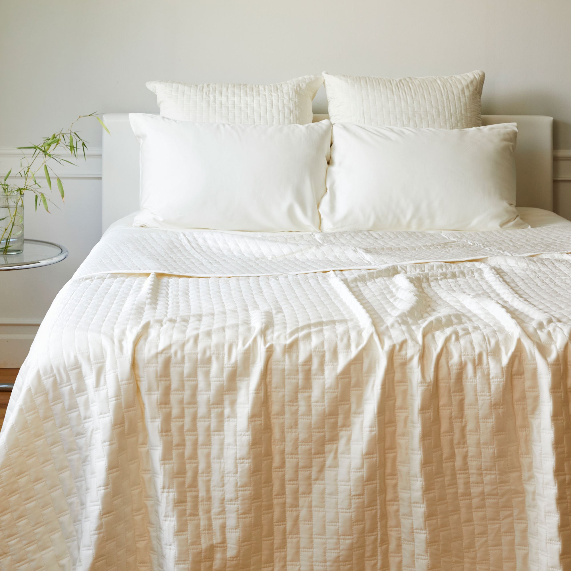 ivory bamboo quilted coverlet and euros on a white headboard and walls with bamboo leaves in a jar nearby