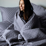 woman wrapped up in dark gray bamboo quilted coverlet and euro shams, smiling