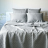 A stone gray bamboo quilted coverlet on a bed, with matching pillows and a plant in the background.