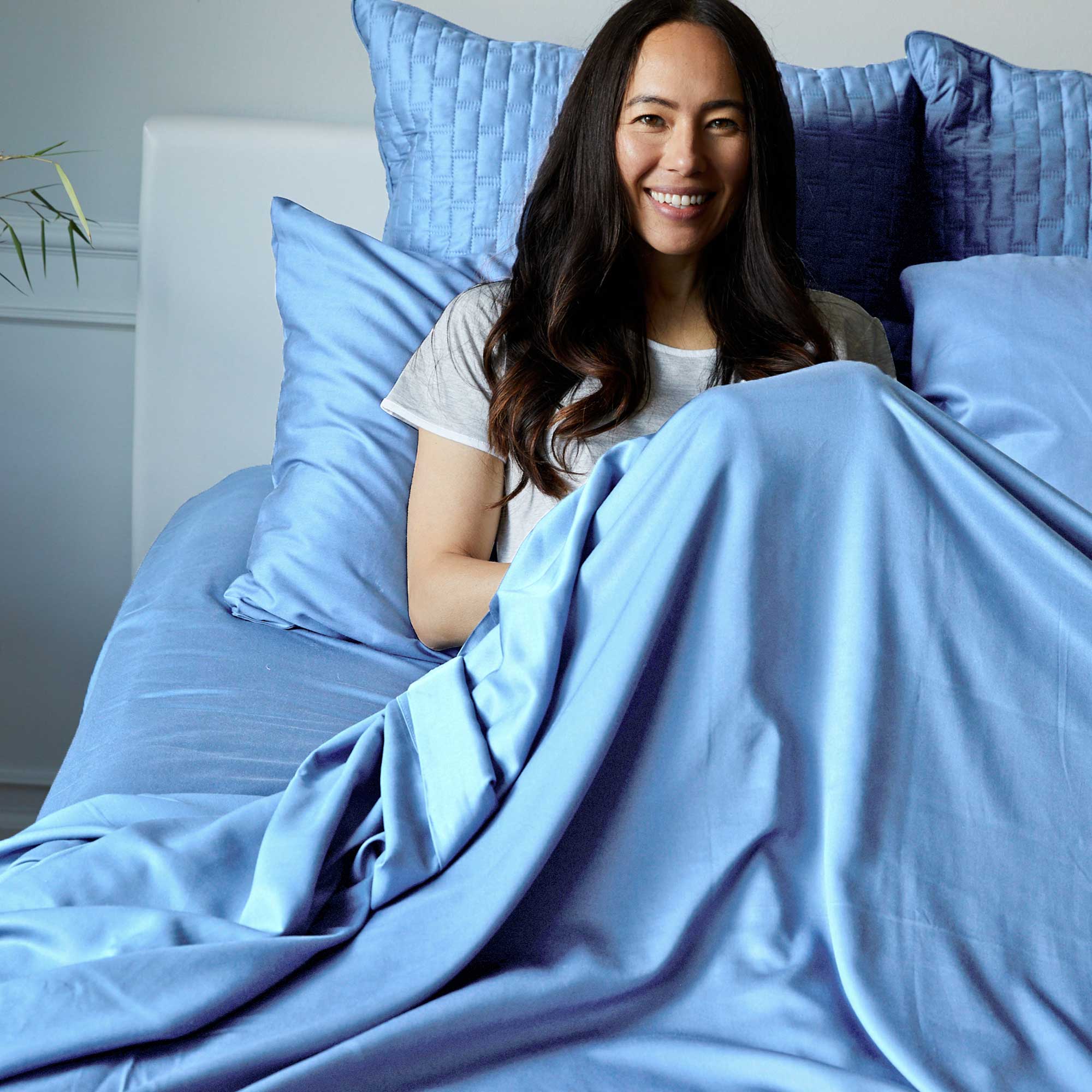 Woman lying in bed with blue bamboo bedding and pillows, smiling.