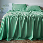 Green bamboo sheets and pillows on a bed in a room with a plant and table.