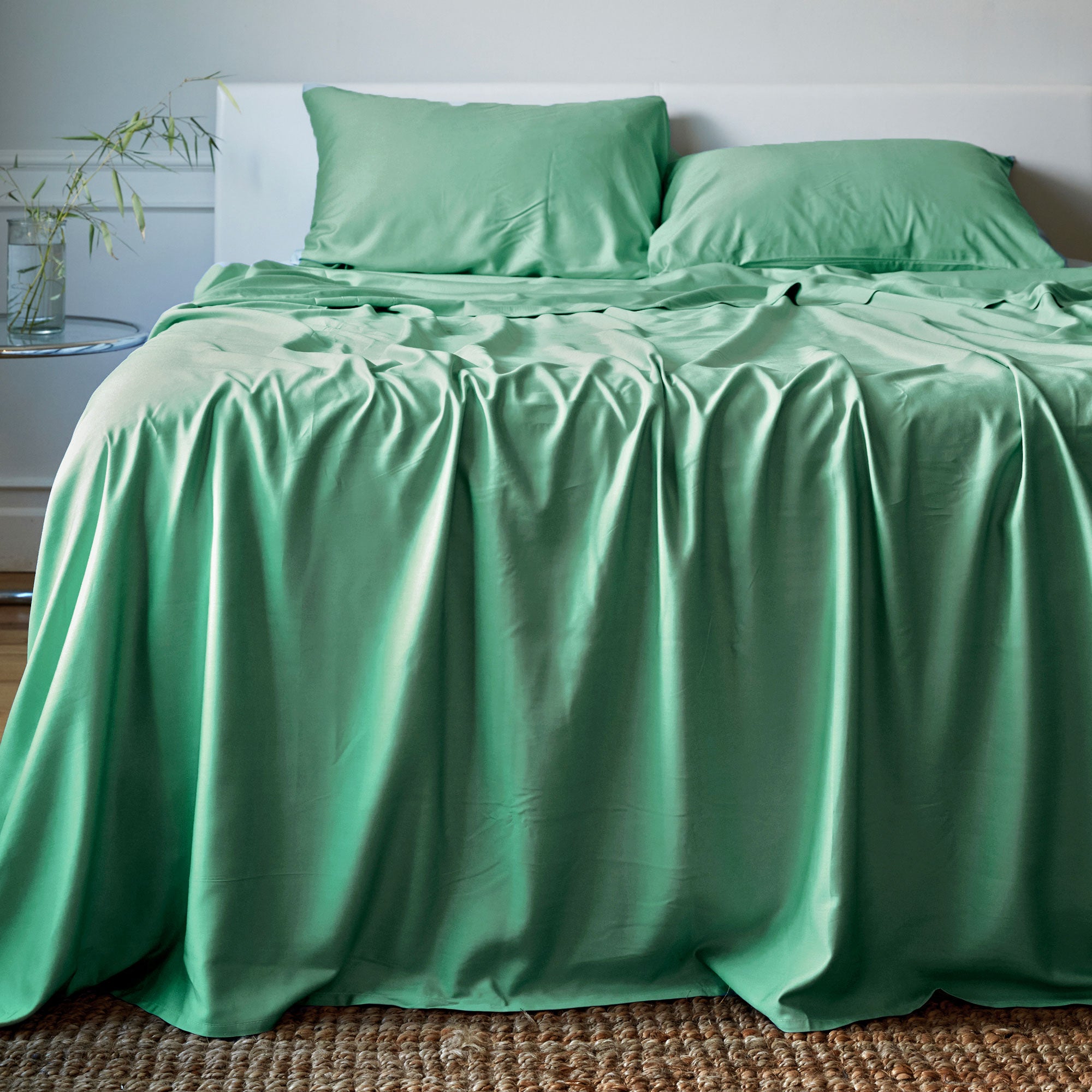 Green bamboo sheets and pillows on a bed in a room with a plant and table.