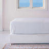 A white fitted sheet on a bed with a window in the background showing a sea view.