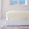 An ivory-colored fitted sheet on a bed with a window in the background showing a beach scene.