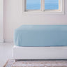 A blue bamboo fitted sheet on a mattress, with a window in the background showing a view of the ocean.