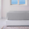 A stone-colored fitted sheet on a bed with a white base, placed in a room with a window overlooking the ocean.
