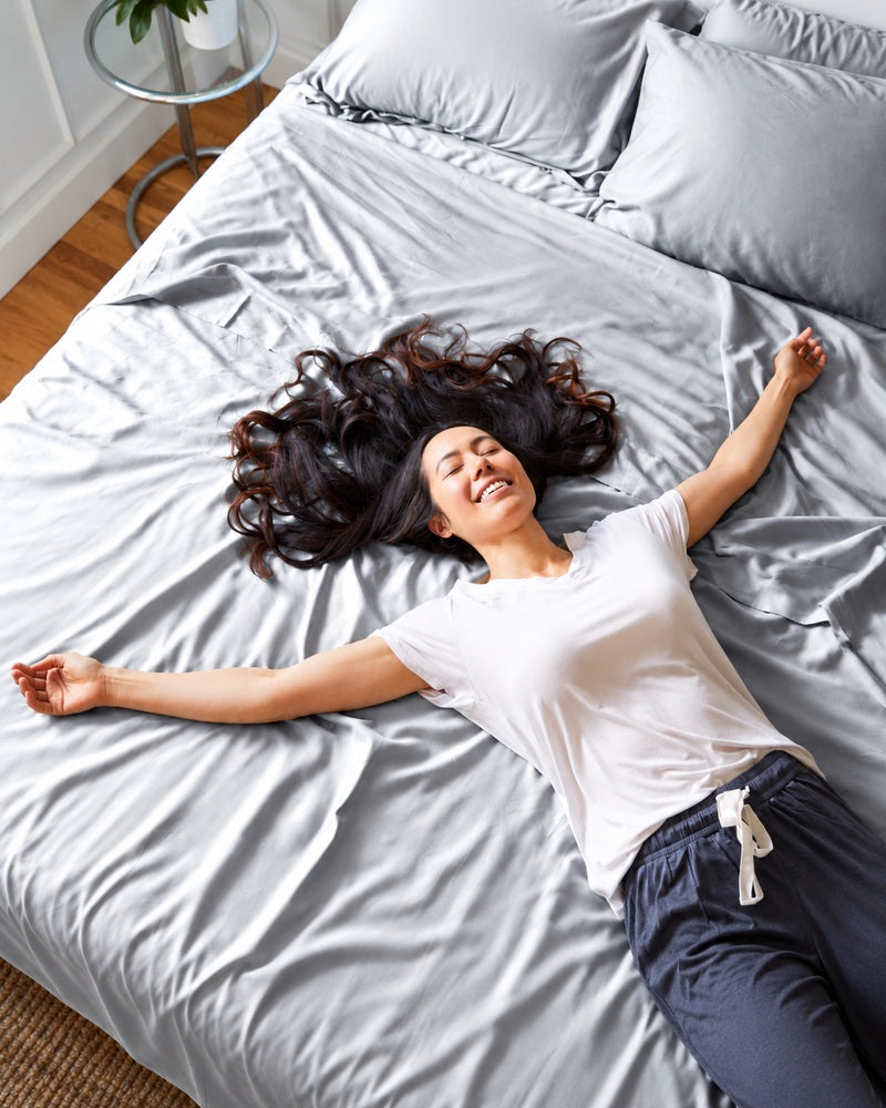 A woman with long dark hair lies stretched out on a bed with light gray satin sheets and matching gray pillows, wearing a white t-shirt and dark navy pants. The photo is taken from a high angle in a bright bedroom.