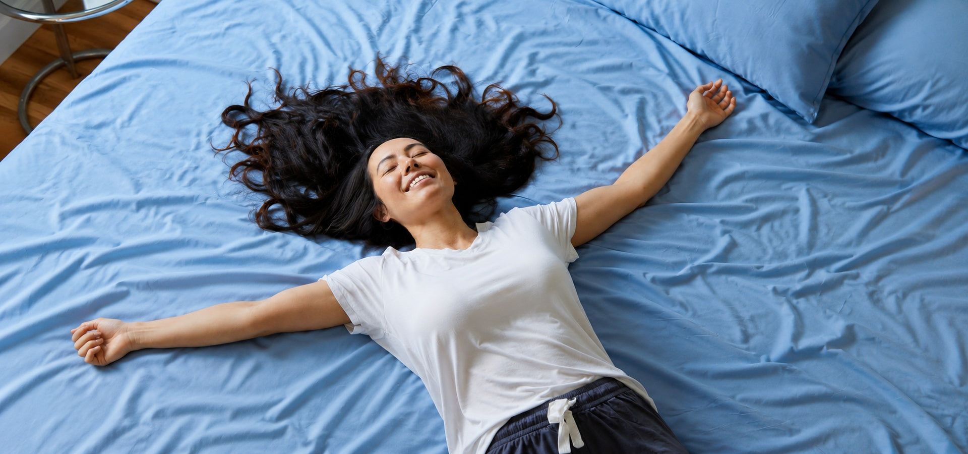 A woman with dark curly hair lies stretched out on a bed with blue sheets and pillows, wearing a white t-shirt and dark shorts.