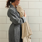 Woman in a gray robe holding a beige towel against a tiled wall.