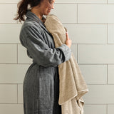 Woman in a gray robe holding a beige towel against a tiled wall.