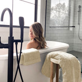 woman sitting in white bathtub with modern white tile walls with beige bamboo towels hanging over tub and stool 