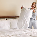 Woman in gray pajamas holding a white bamboo pillowcase over a bed with white bedding.