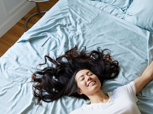 woman with dark hair happy laying on light blue bamboo sheets on a bed